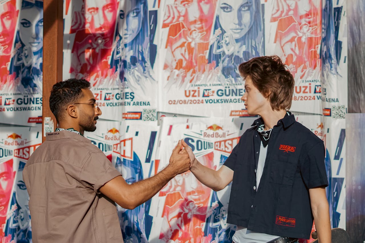Two young men sharing a handshake in front of concert posters in a vibrant street setting.